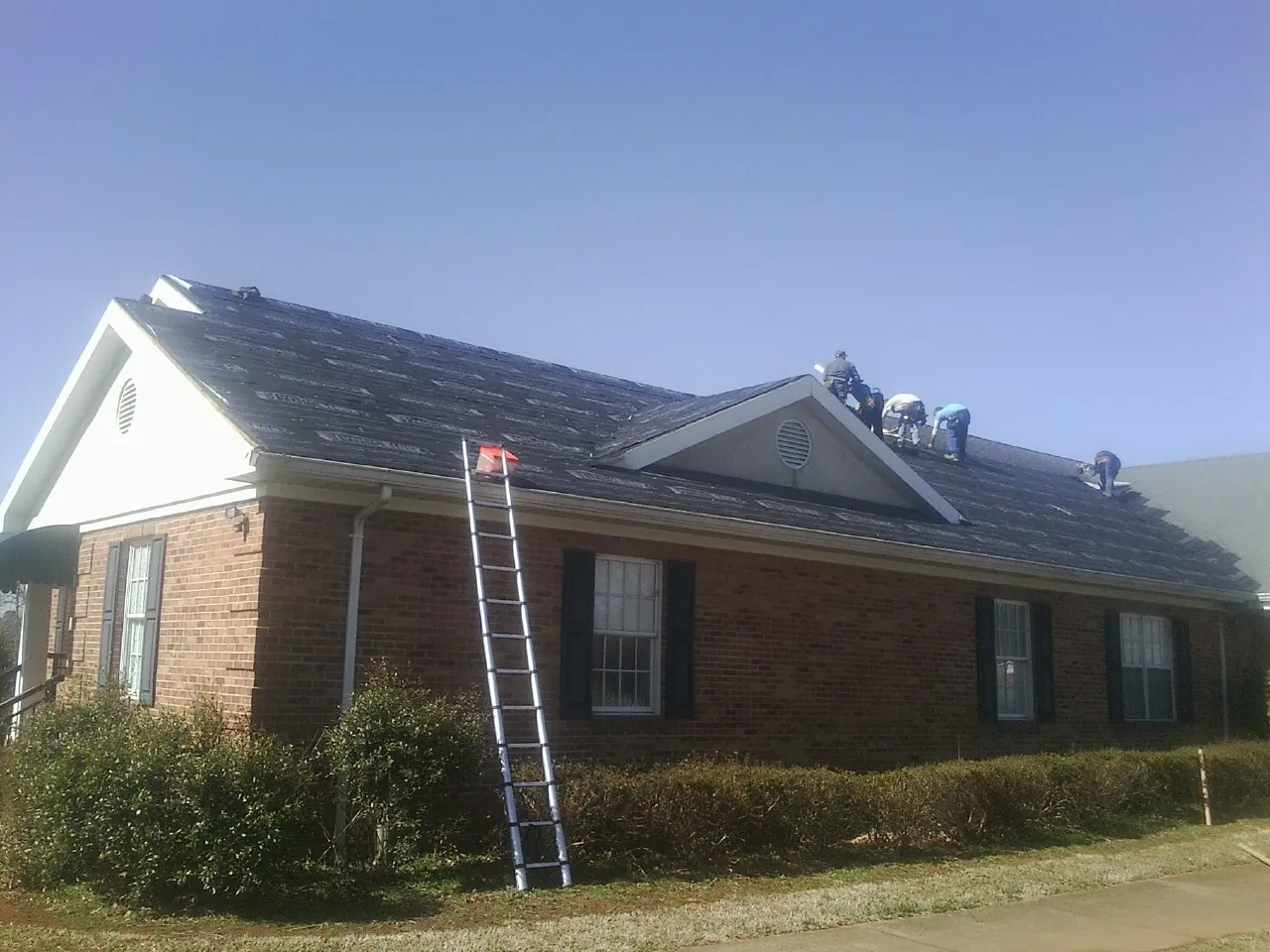 Crew tearing shingles off a brick ranch roof with a ladder set — Metal Roof Repair work by our crew in New Ellenton