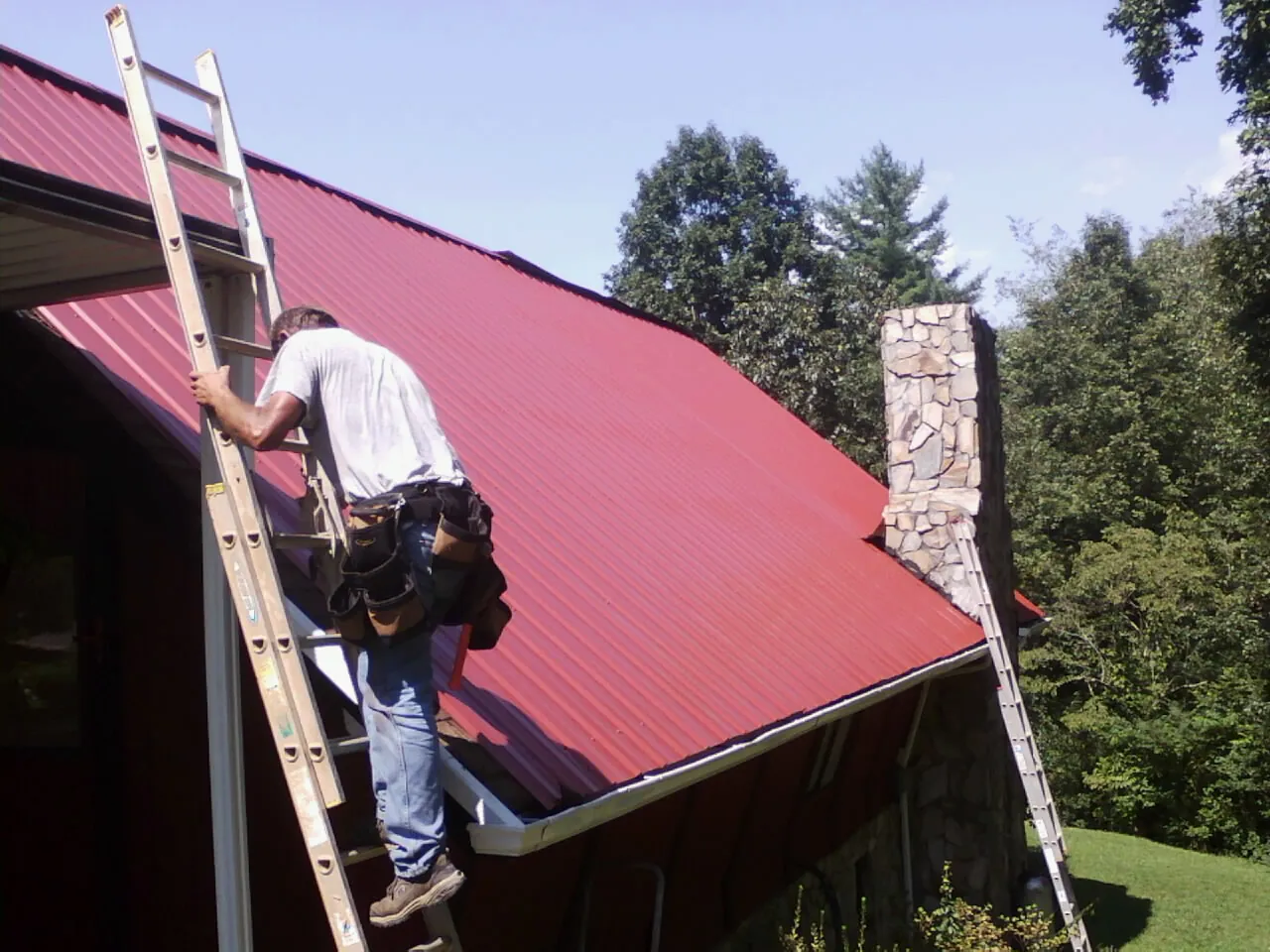 Roofer on a ladder beside a red metal roof and stone chimney — Soffit & Fascia Repair by Roofing Brothers in New Ellenton